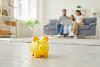 Salvadanaio giallo a maialino in primo piano, con sullo sfondo una famiglia seduta sul divano di casa, sfocata.